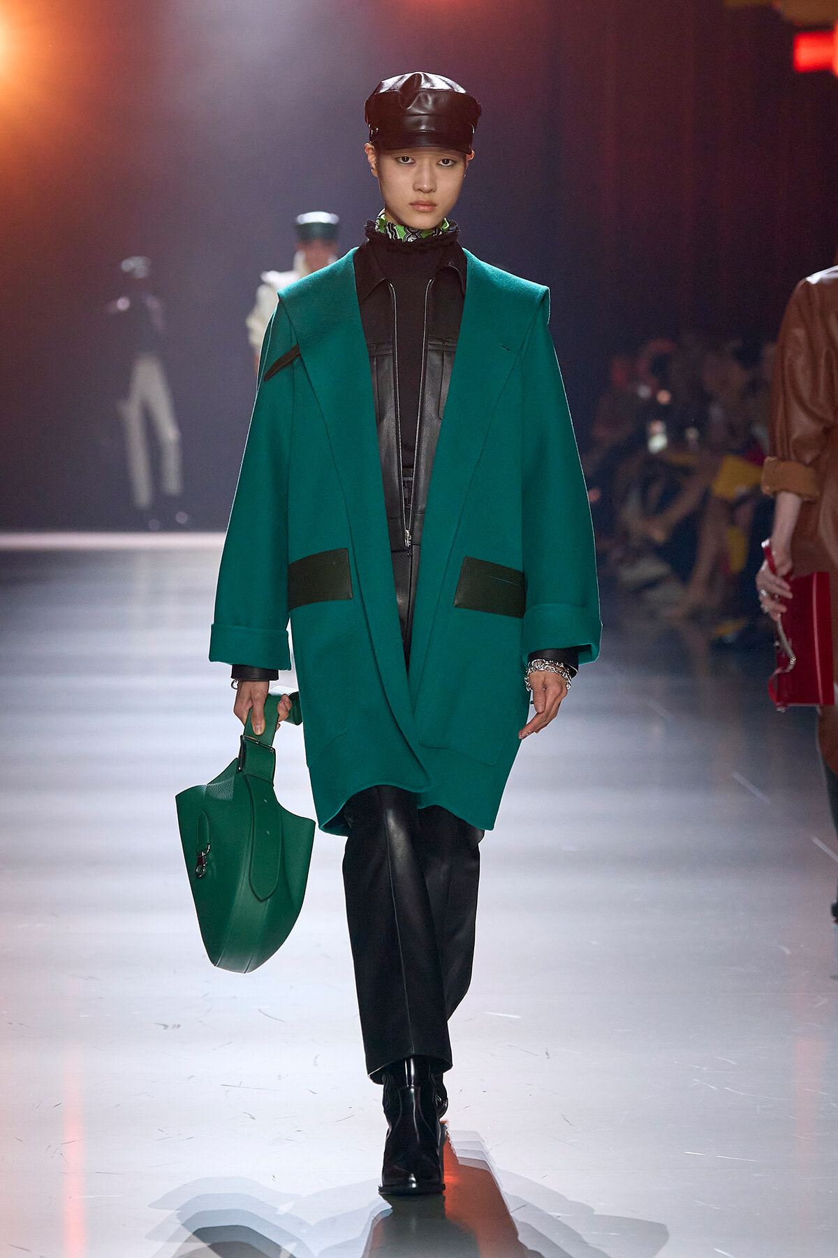 Model on a runway wearing an emerald green oversized coat over a black leather outfit, black cap, and matching green handbag.