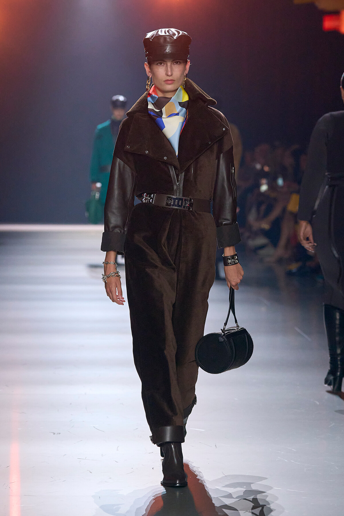 Model walking a runway in a dark brown corduroy jumpsuit with leather sleeves, wide belt, black cap, colorful scarf, and a round black handbag.