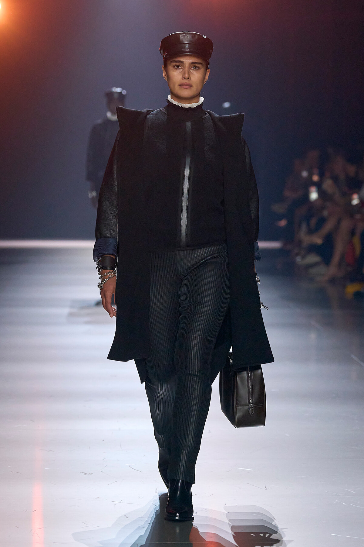 Female model walks a fashion runway in a black, oversized coat with a cap, pinstriped pants, and a dark handbag understage lighting.