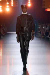 Model walks a fashion runway in an all-black leather outfit with a cap and white scarf under warm stage lights.