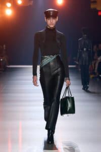 Model walking the runway in an all-black outfit: turtleneck, leather pants, bandana belt, black cap, and a black structured handbag.