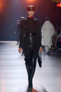 Female model walking runway in black turtleneck, high-waisted leather pants, belt, knee-high boots, and a black cap, holding a black bag.