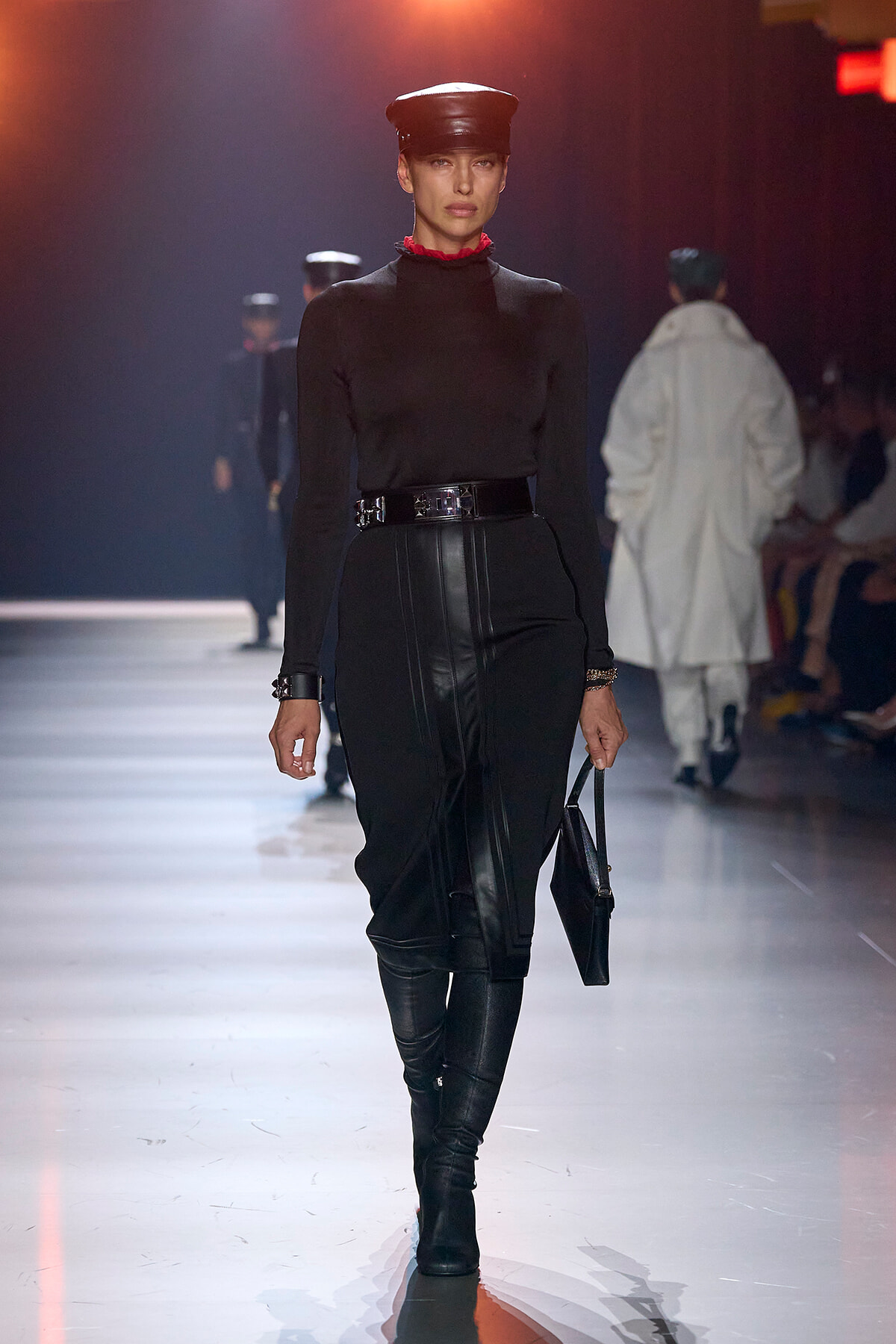Female model walking runway in black turtleneck, high-waisted leather pants, belt, knee-high boots, and a black cap, holding a black bag.