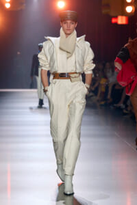 Model walks the runway in an all-white quilted jumpsuit with a beige scarf, brown belt, and white ankle boots under warm stage lights