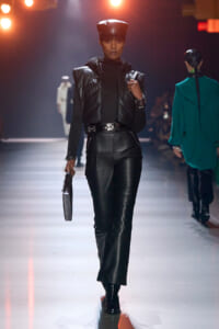 Model walking the runway in a black leather outfit with a padded vest, cap, and matching boots, holding a small silver bag.
