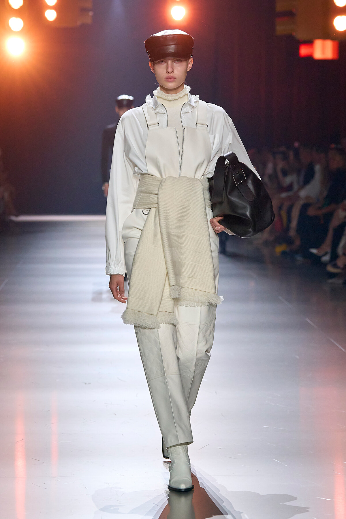 Model on a runway wearing an all-white ensemble: oversized zip-front top, quilted pants, a wide beige scarf belted at the waist, cream turtleneck, black cap, and a black bag on the arm.