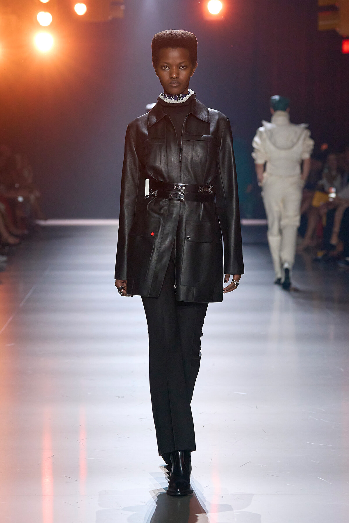 Model walks a fashion show in a black leather belted coat, matching black pants and boots under warm stage lights.