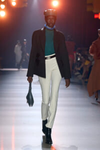Model walks the runway in a teal turtleneck, black blazer, white pants, and black boots, with a beret and blue scarf, carrying a small dark handbag.