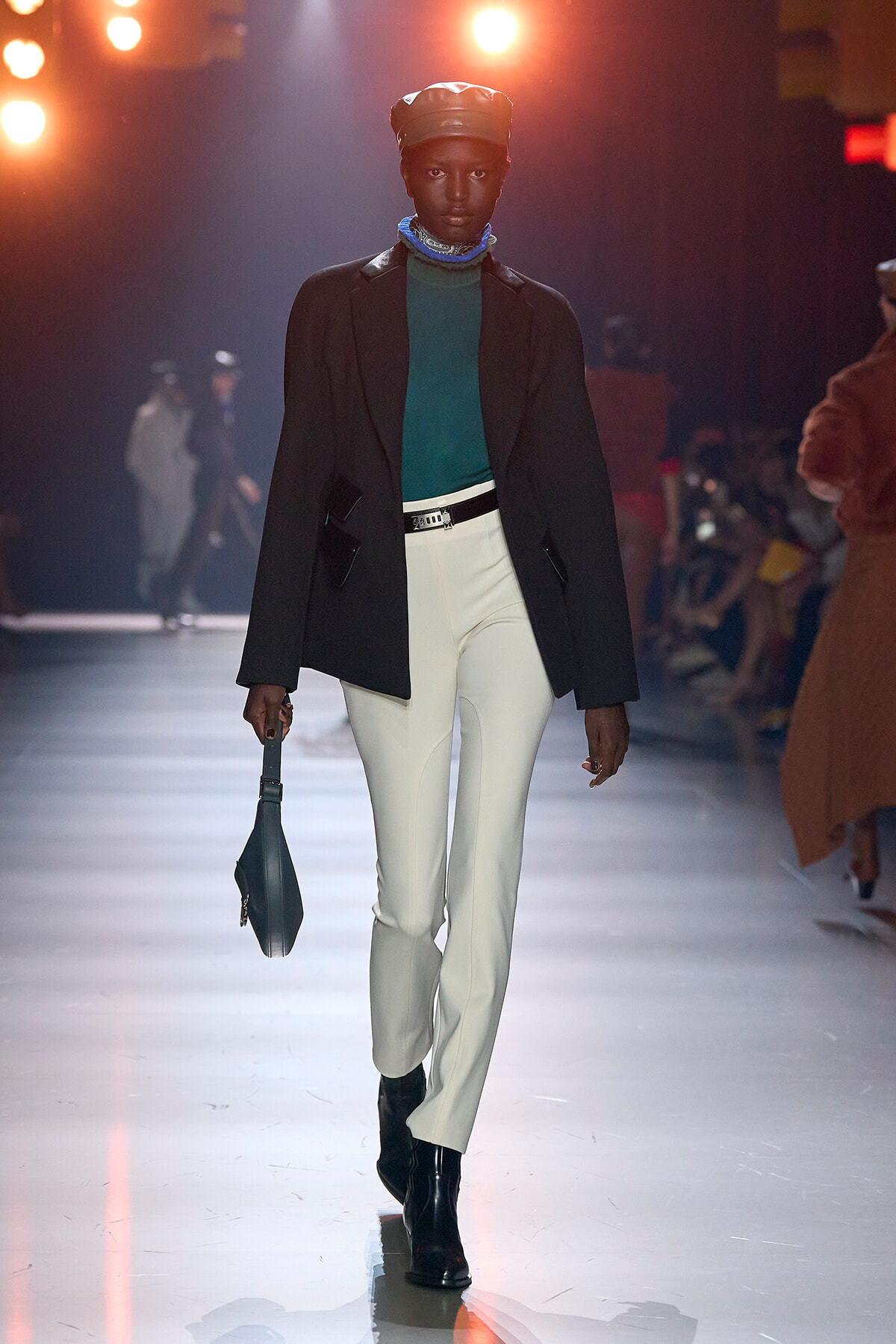 Model walks the runway in a teal turtleneck, black blazer, white pants, and black boots, with a beret and blue scarf, carrying a small dark handbag.