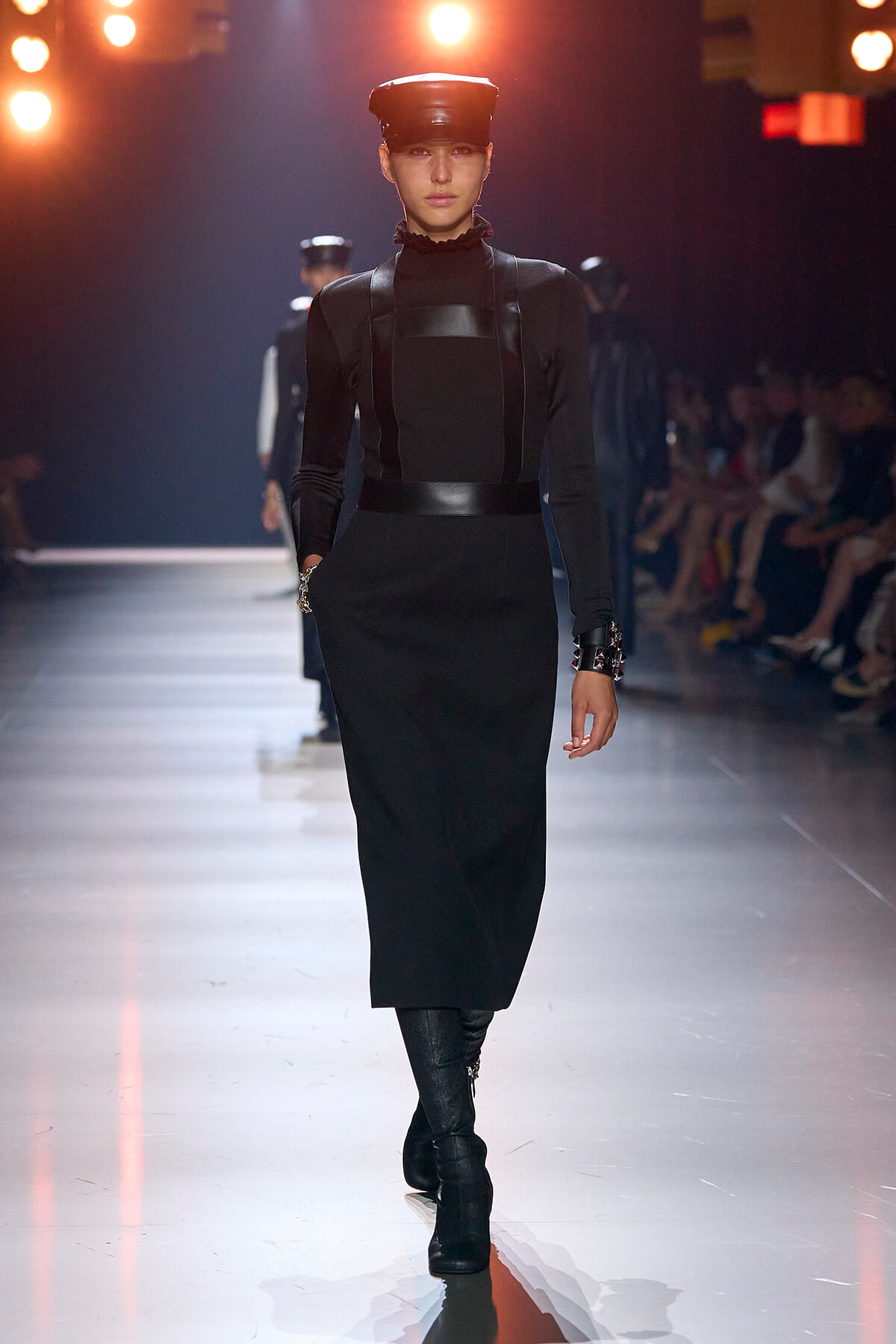 Model walks the runway in a black, high-neck dress with leather accents and a matching cap; bright stage lights in the background.