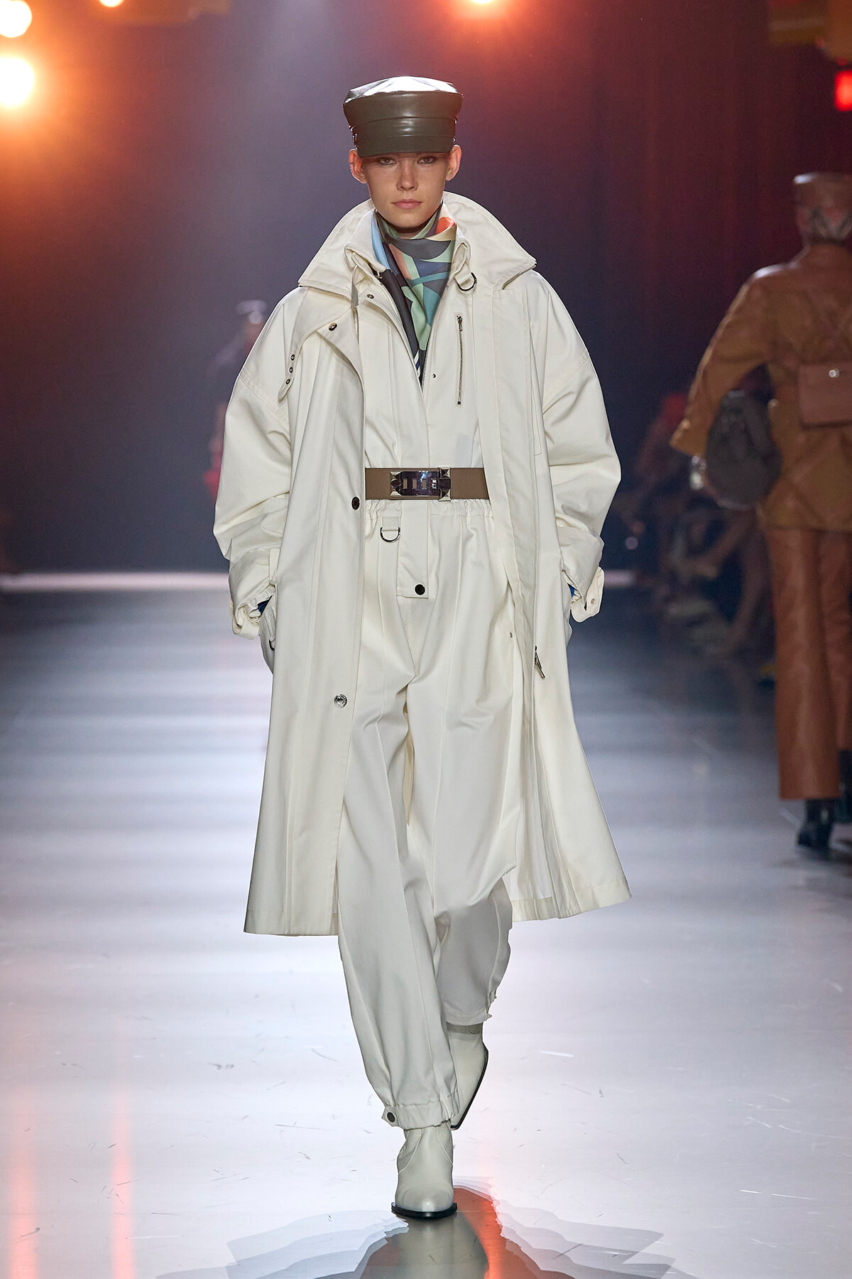Model walking a runway in an oversized white trench coat with a brown belt, matching white pants, a black cap, and a blue-green scarf at the neck.