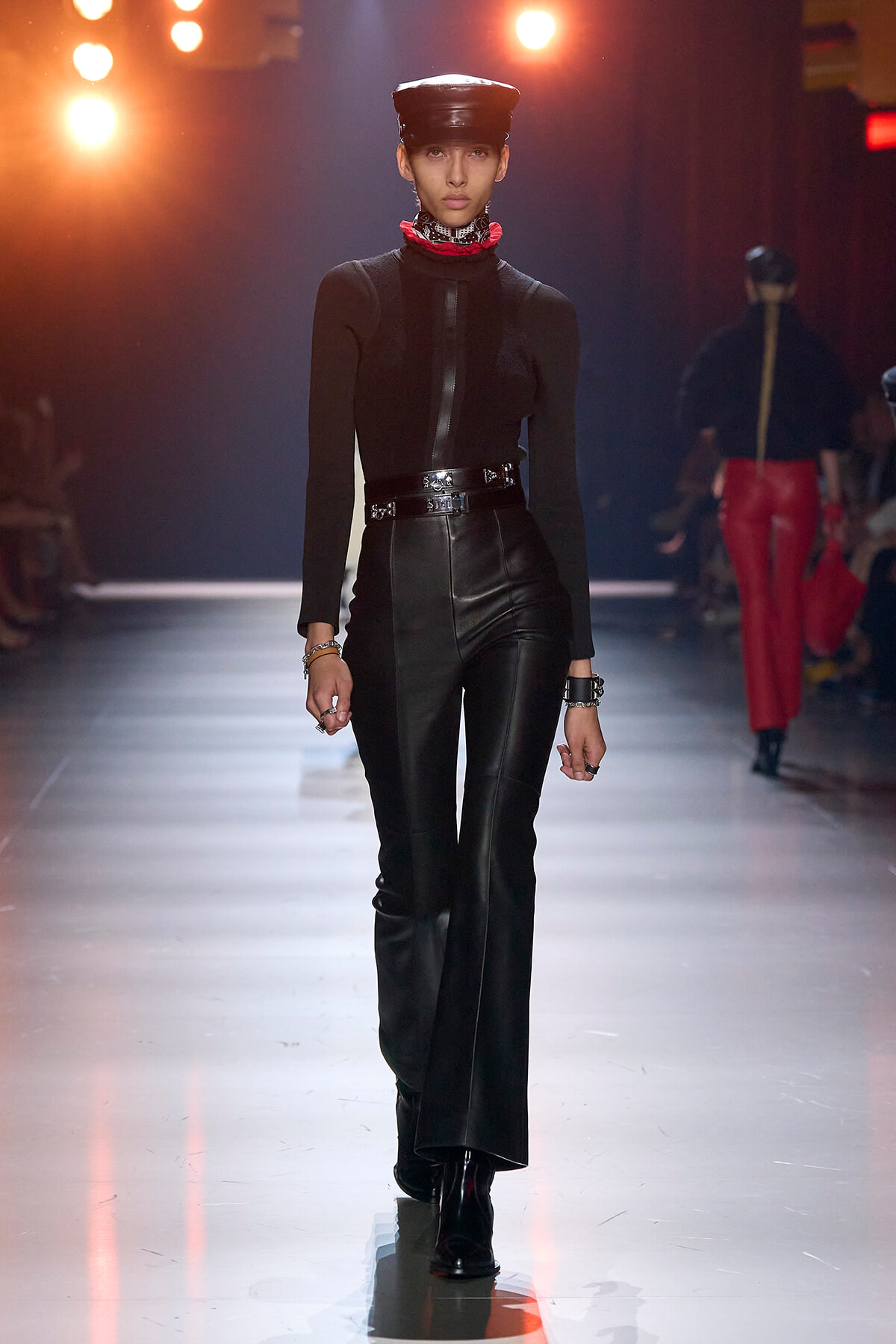 Model walking a runway in a black leather outfit with a cap, red neck scarf, and silver bracelets under bright stage lights.