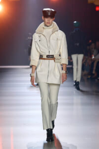 Model walks a fashion runway wearing a white belted coat with a high collar, cream turtleneck, matching pants, black cap, and black boots.