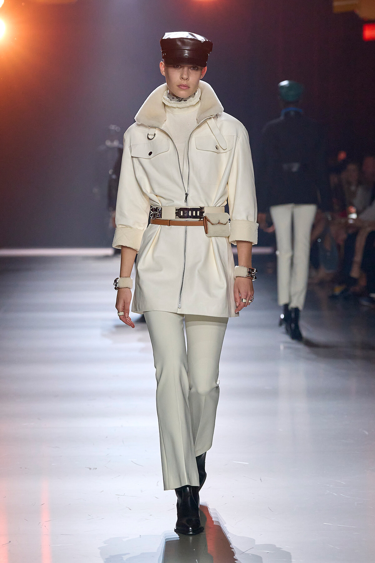 Model walks a fashion runway wearing a white belted coat with a high collar, cream turtleneck, matching pants, black cap, and black boots.