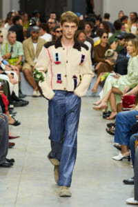 Male model walks a runway in a cream knit cardigan with colorful patch motifs and blue wide-leg jeans.