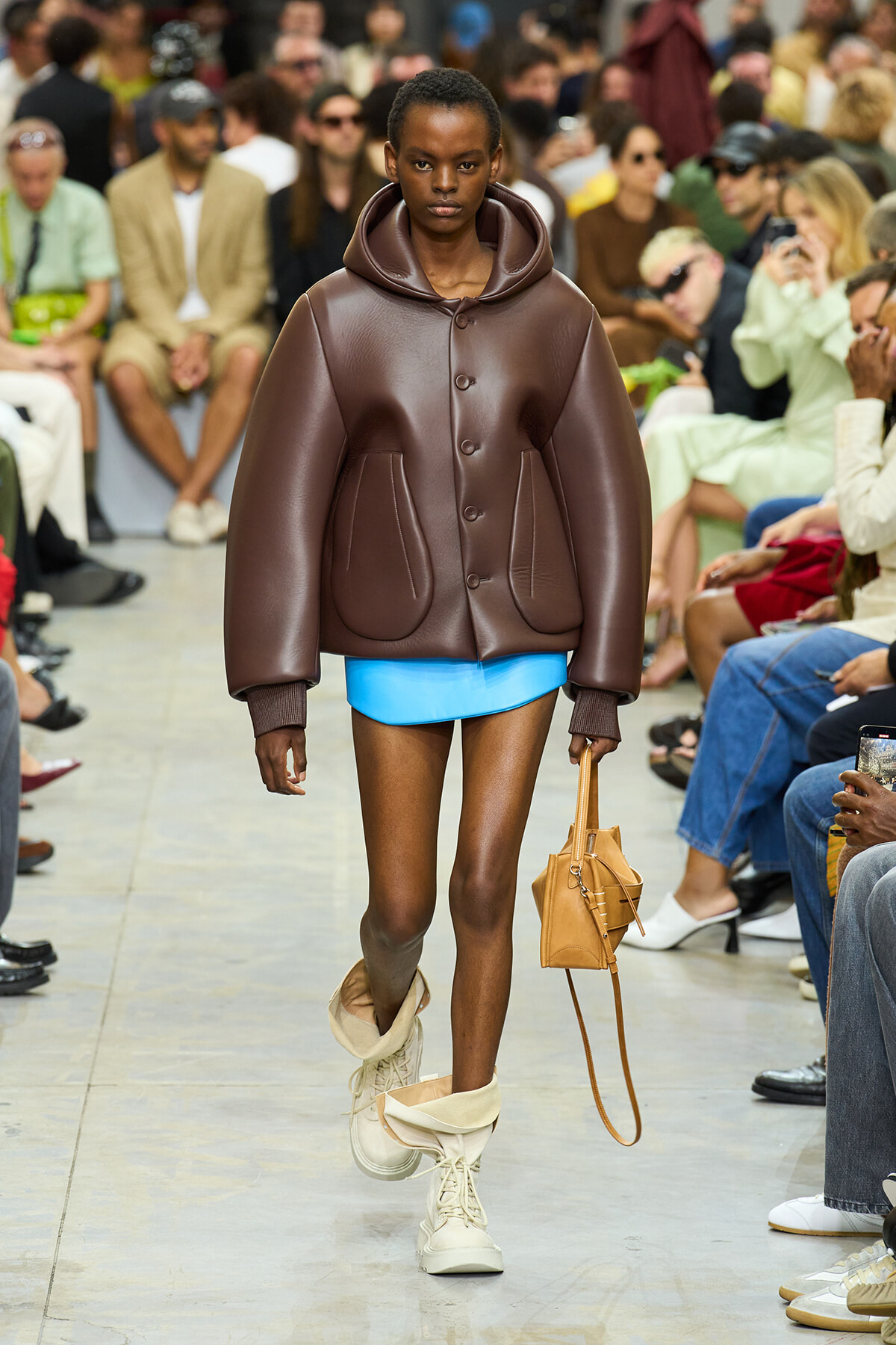 Model walking a runway in an oversized brown leather jacket, bright blue shorts, cream combat boots, and a tan handbag.