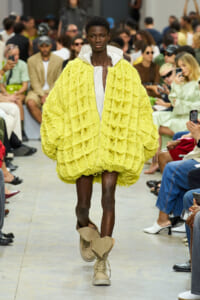 Runway model strides toward the camera in a bright yellow, oversized textured coat with beige boots, audience in background.