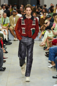Male model walks a runway in a red textured cardigan with white window-like patterns, dark collar, paired with gray jeans and beige sneakers.