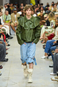 Male model walks the runway in a green oversized structured jacket, light blue rolled denim shorts, and beige boots at a fashion show.