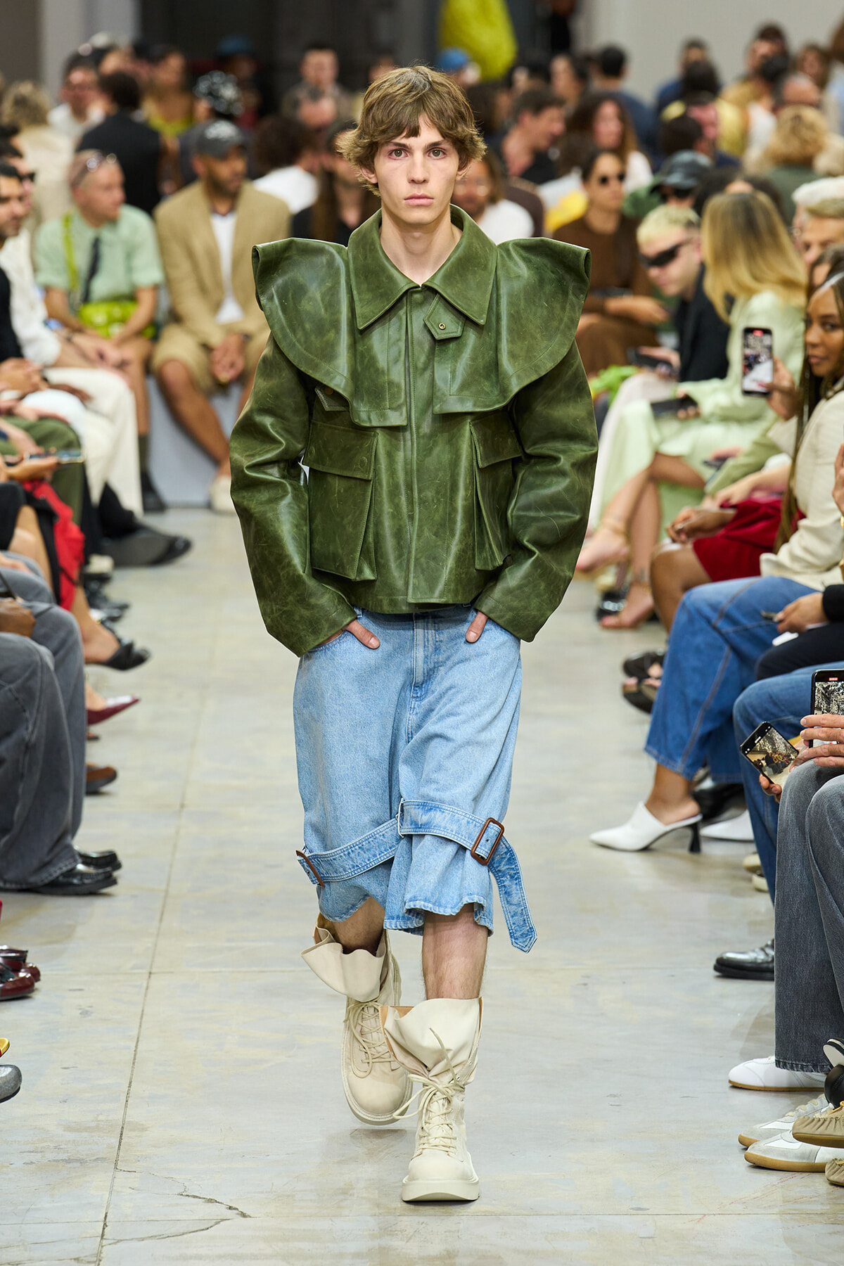Male model walks the runway in a green oversized structured jacket, light blue rolled denim shorts, and beige boots at a fashion show.
