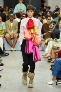 Model walks the runway wearing a pale pink shirt with oversized multicolored fabric sculpted across the chest, black pants, and tan buckled boots.