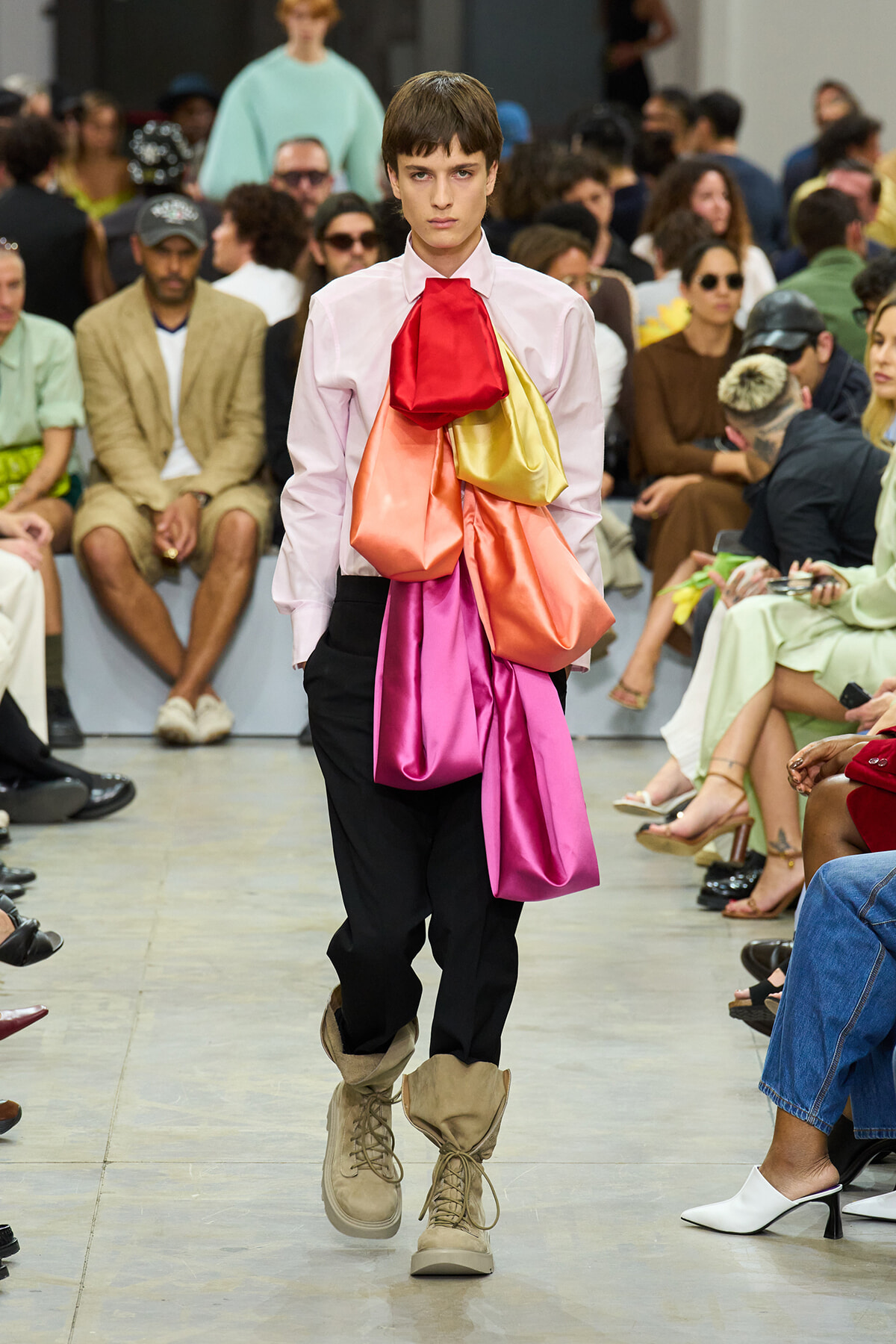 Model walks the runway wearing a pale pink shirt with oversized multicolored fabric sculpted across the chest, black pants, and tan buckled boots.