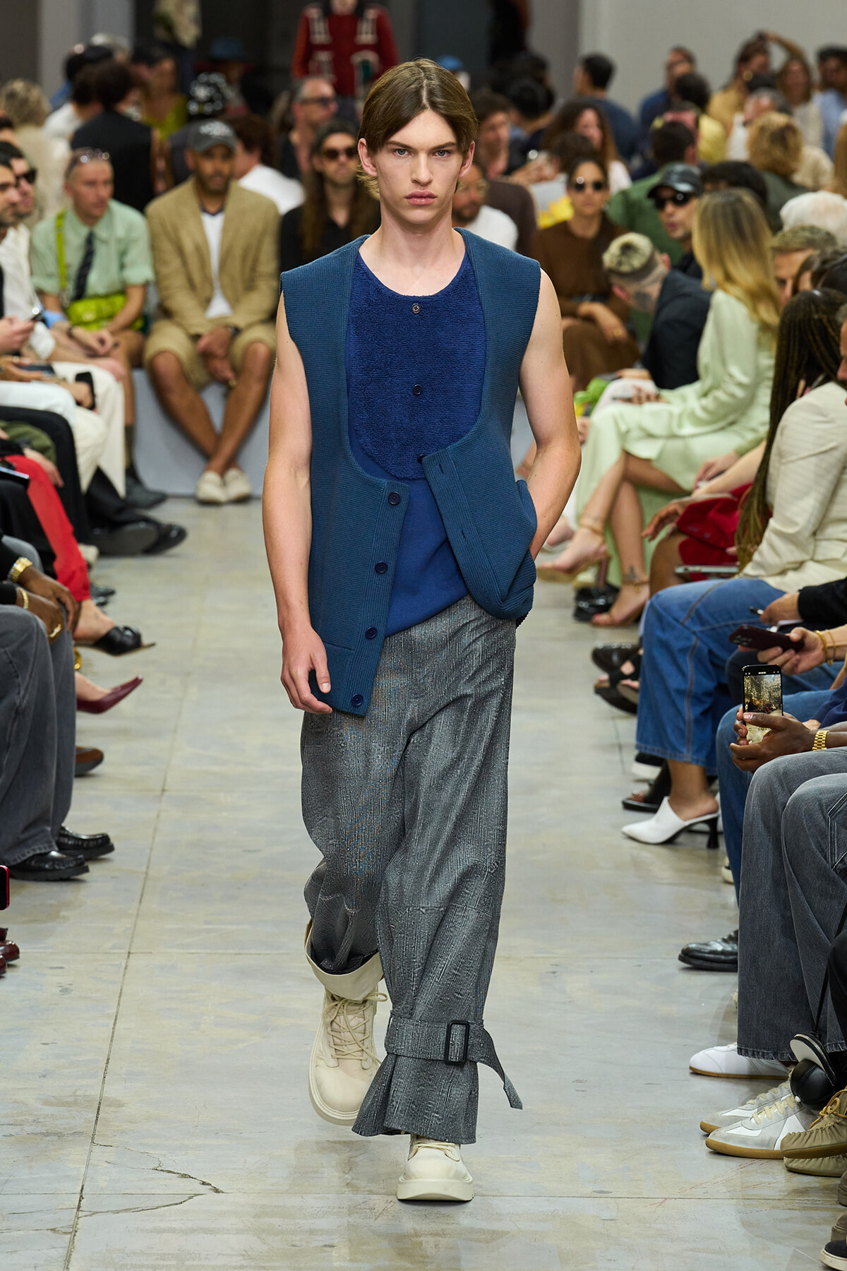 Male model walks a runway wearing a blue sleeveless vest over a blue knit top and wide gray trousers; audience watches from both sides.