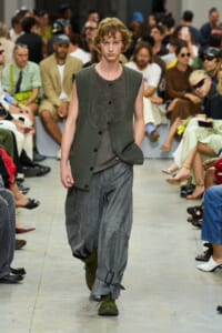 Male model walks a runway in loose gray textured pants and an olive-green sleeveless cardigan, audience seated along the sides behind him.