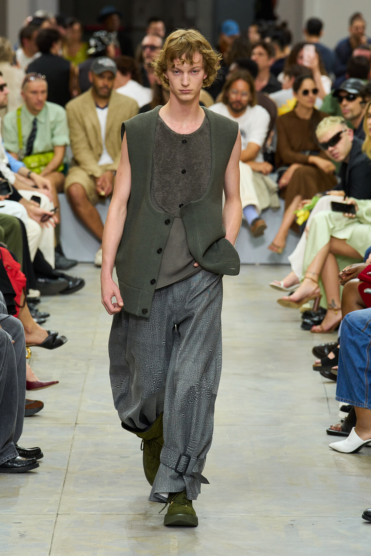 Male model walks a runway in loose gray textured pants and an olive-green sleeveless cardigan, audience seated along the sides behind him.