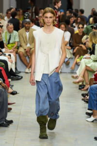 Model on a runway wearing a white sleeveless vest over a white top with loose blue denim pants and green combat boots.