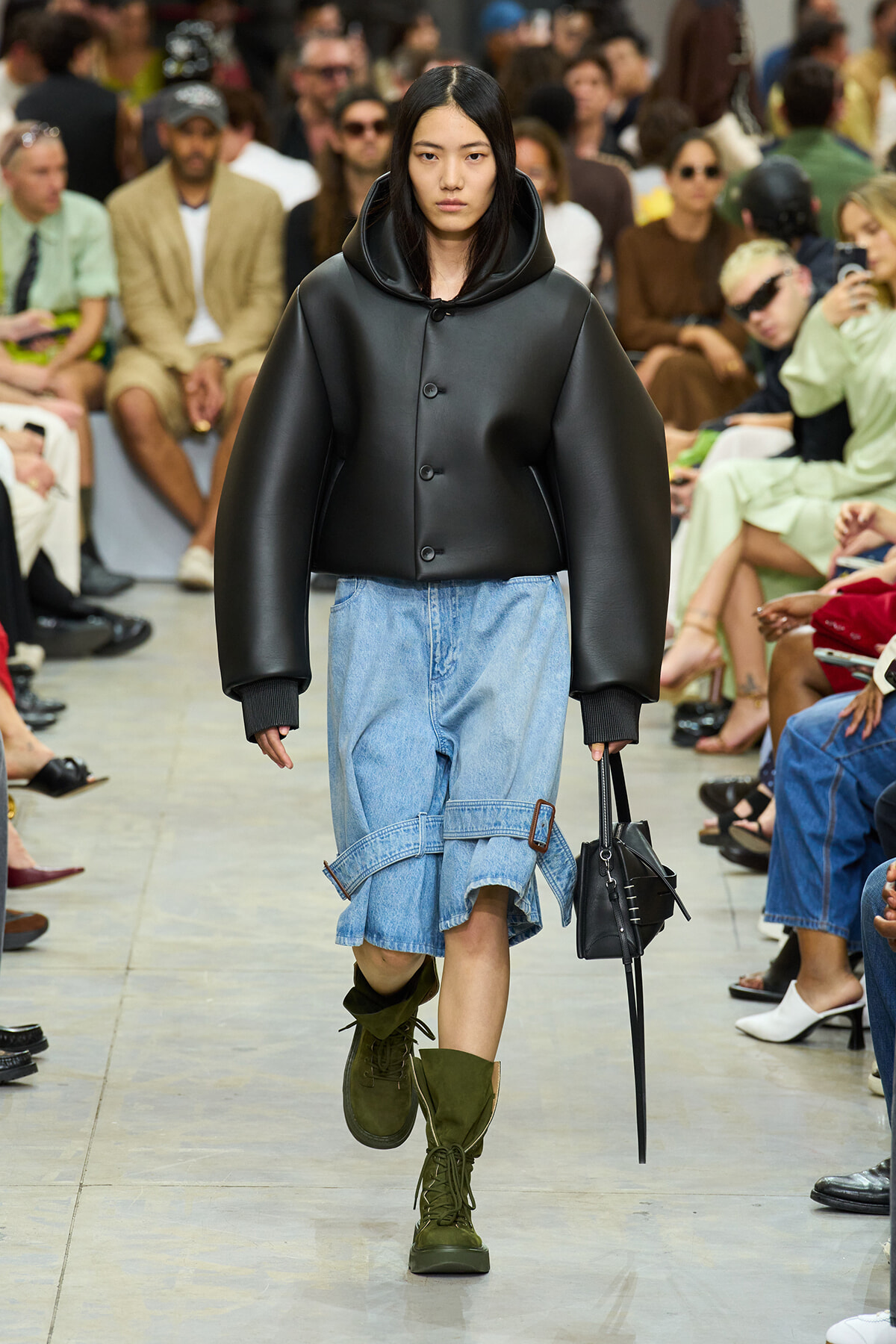 Model walks the runway in a oversized black leather jacket with a hood, paired with a light-blue denim skirt and olive combat boots.