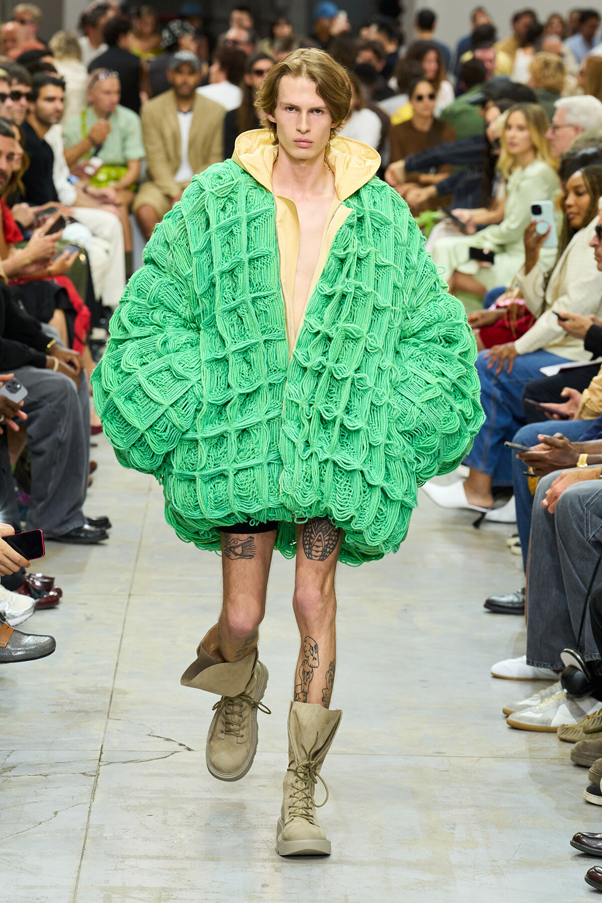 Male model walks a fashion show wearing a large green knitted coat with a tan collar and beige lining, audience in background.