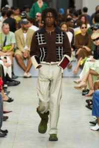 Male model walks the runway in a chunky brown cardigan with white grid patterns, beige wide-legged pants, and green boots as spectators watch.