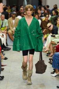 Model walking a fashion runway in a bright green oversized top, black shorts, tan boots, and carrying a brown tote bag; audience in blurred background.