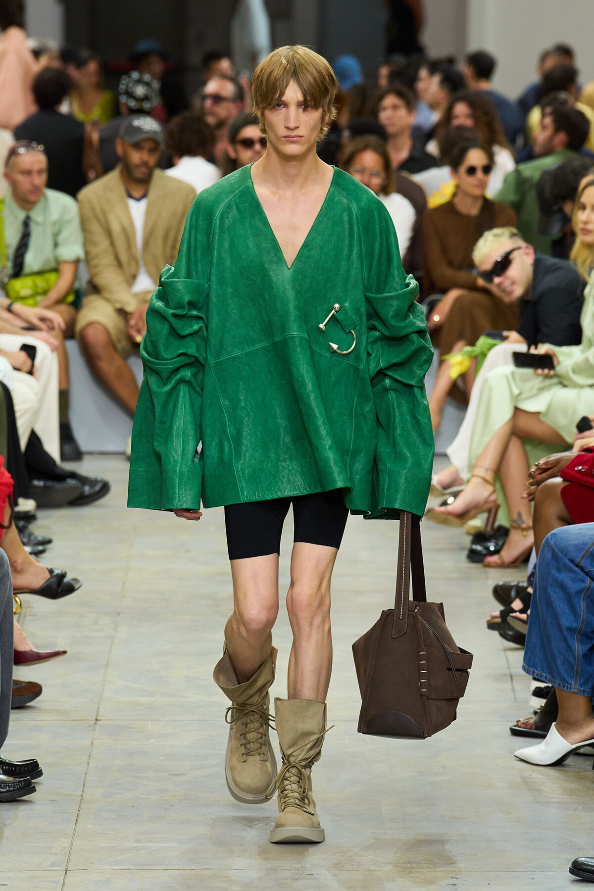 Model walking a fashion runway in a bright green oversized top, black shorts, tan boots, and carrying a brown tote bag; audience in blurred background.