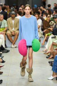 Model on a fashion runways wearing a light blue oversized sweater and tan combat boots, with large pink and green circular cushion-like accessories hanging at the hips as an avant‑garde detail.