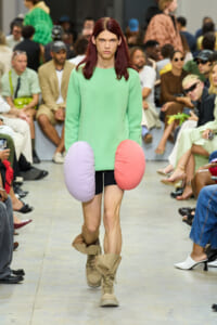Model on a runway wearing a mint green oversized sweater and large pastel hip cushions (pink and lavender) with tan combat boots, audience in background.
