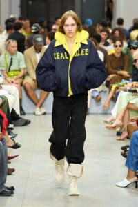 Runway model in an oversized navy jacket with a neon yellow collar, black pants, and cream boots as spectators watch.