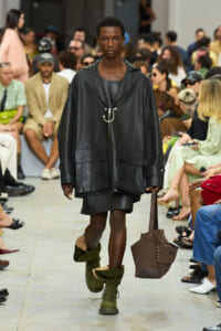 Model walks a runway in an oversized black leather jacket with large pockets, olive-green boots, and a brown tote bag.