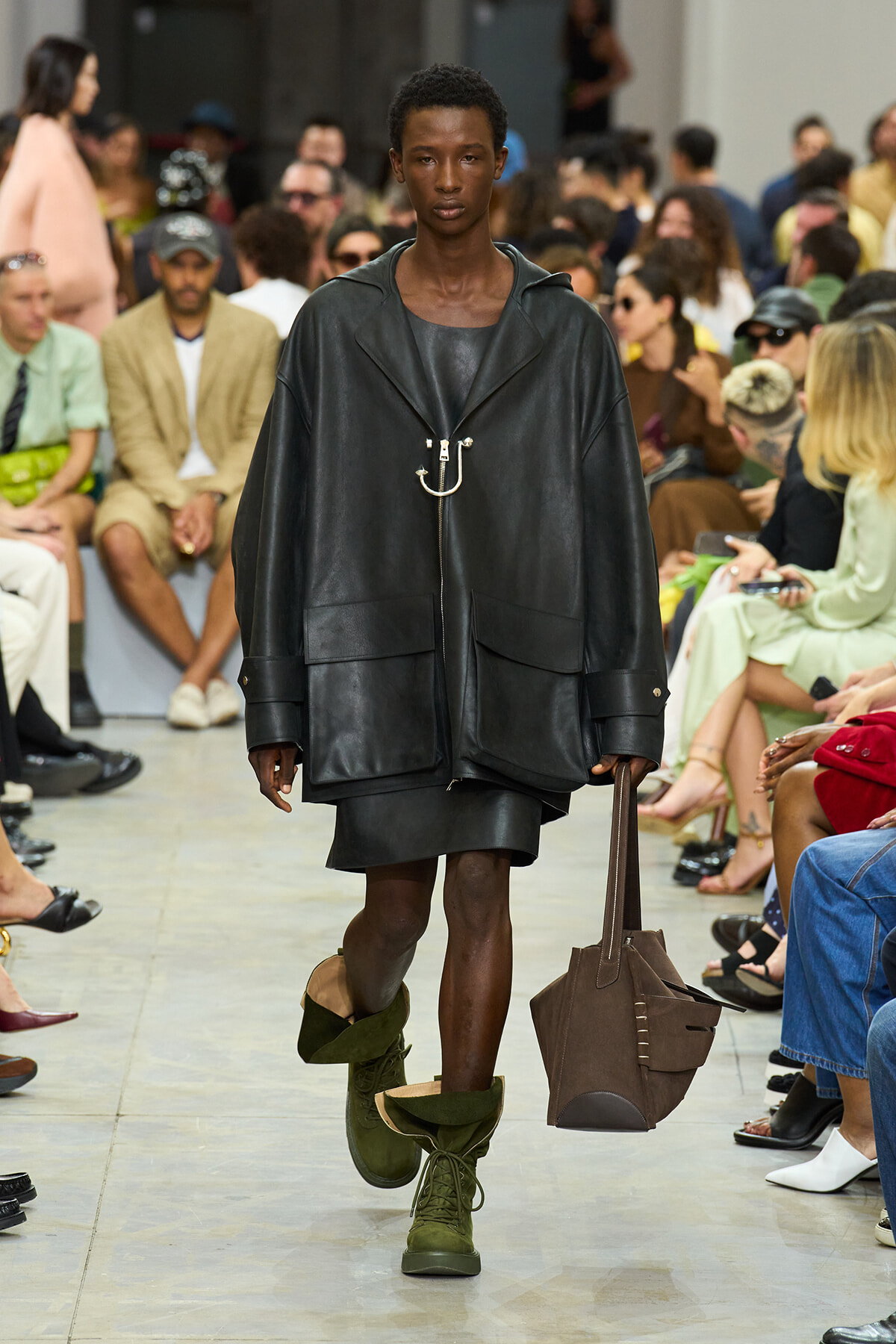 Model walks a runway in an oversized black leather jacket with large pockets, olive-green boots, and a brown tote bag.