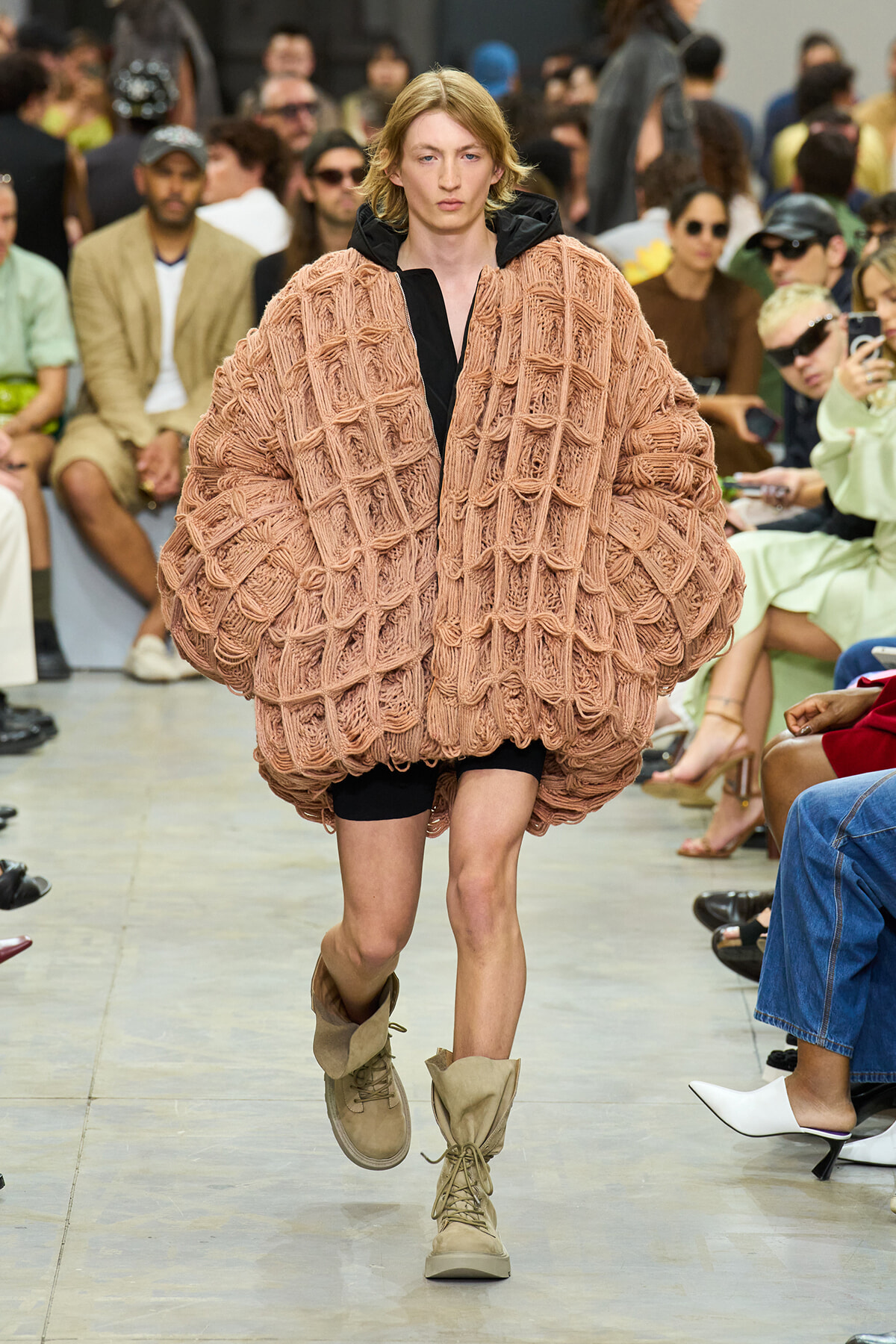 Model walks the runway in an oversized peach knit poncho with a black outfit underneath and beige lace-up boots.