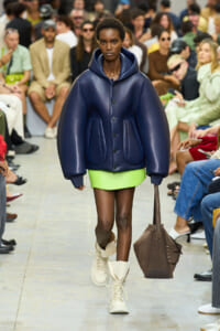 Model walking a fashion runway in an oversized navy puffer jacket, bright lime mini skirt, and white boots, carrying a brown handbag.