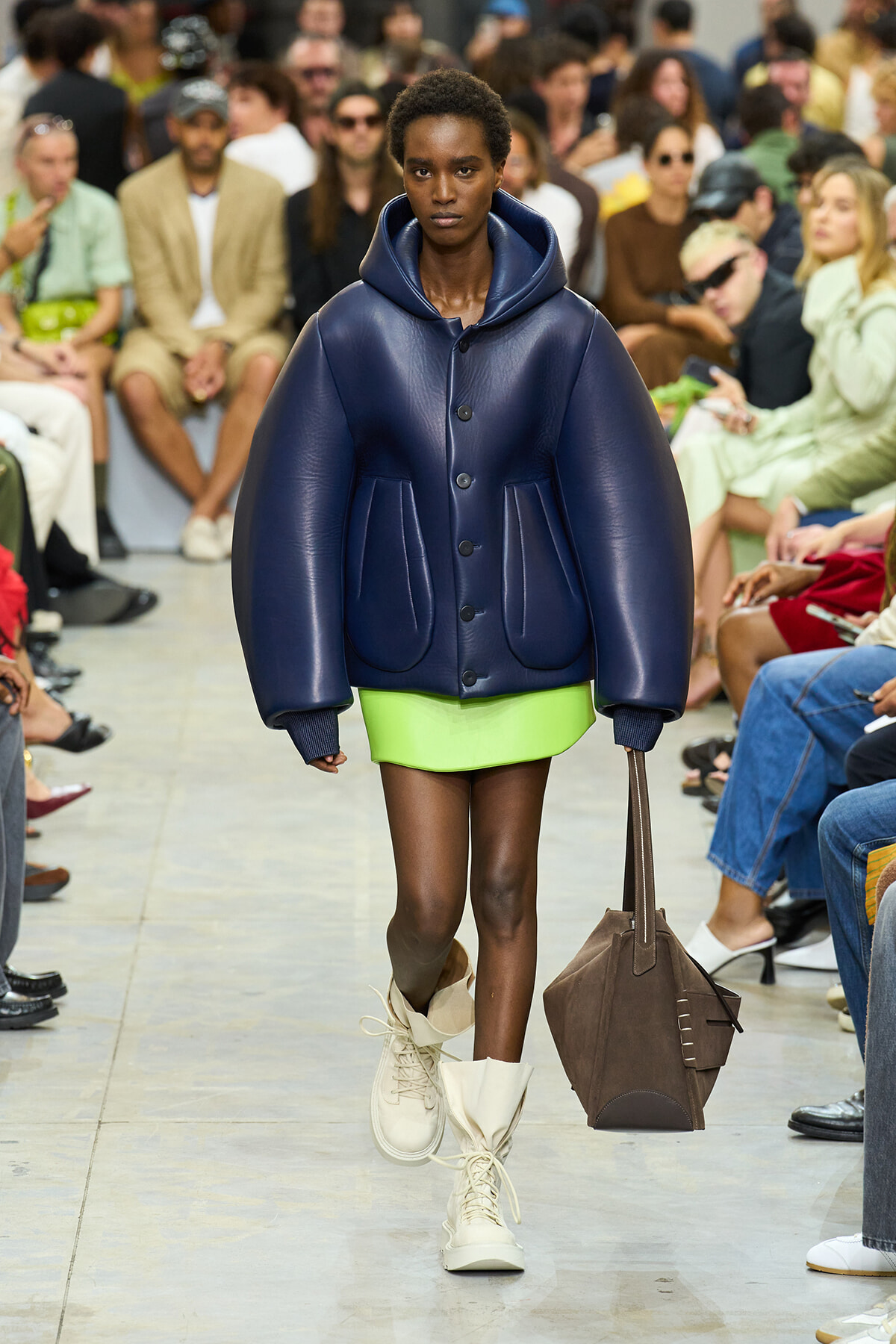 Model walking a fashion runway in an oversized navy puffer jacket, bright lime mini skirt, and white boots, carrying a brown handbag.