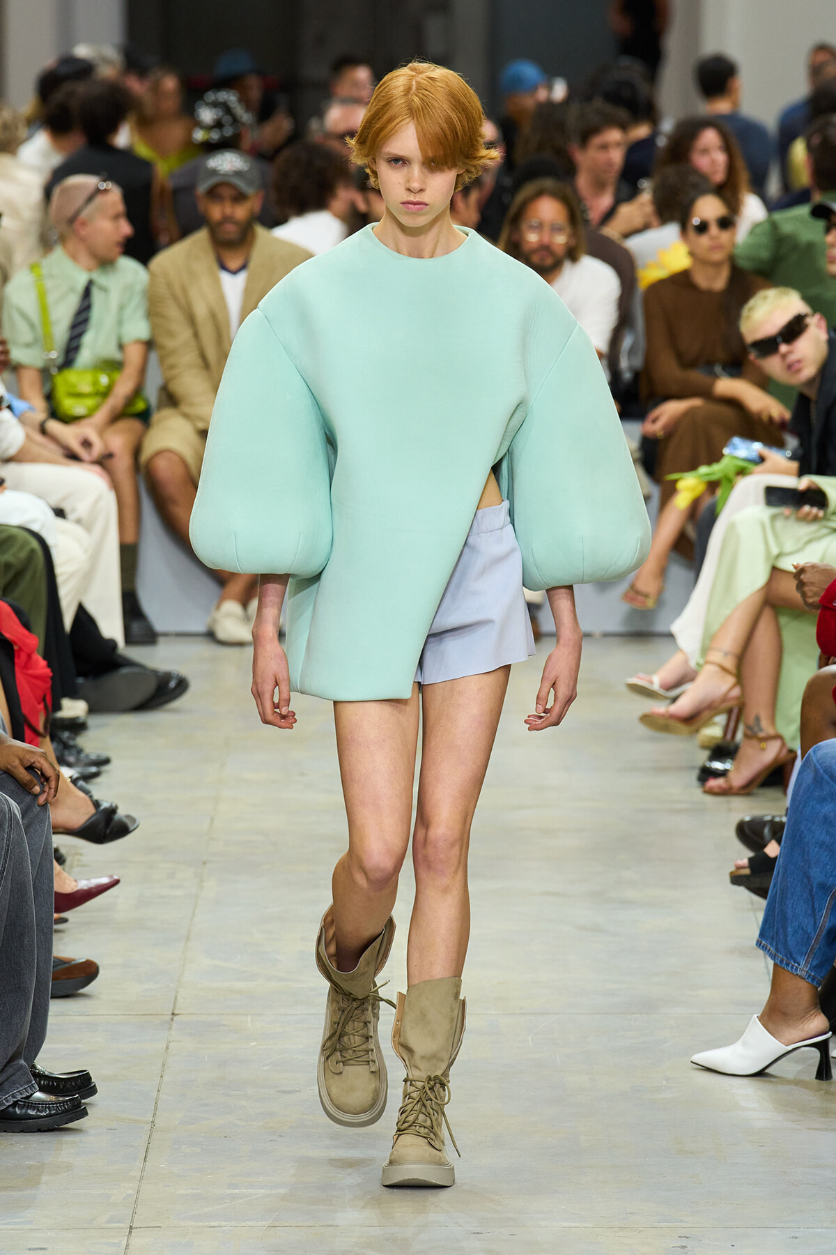 Model walks a fashion runway in an oversized mint green top with voluminous sleeves, pale blue shorts, and tan combat boots; audience blurred in the background.