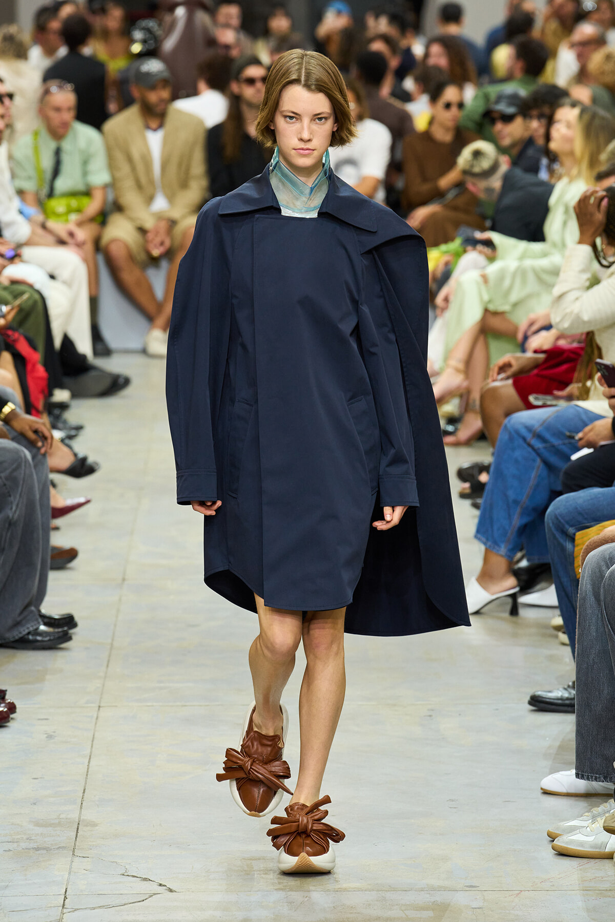 Model walks the runway in a navy oversized cape-like coat and brown bow-front sandals as a fashion show audience watches closely.