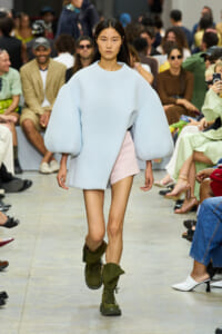 Model walks the runway in a pale blue oversized top with voluminous sleeves, paired with a pale pink mini skirt and olive green combat boots, audience in background.