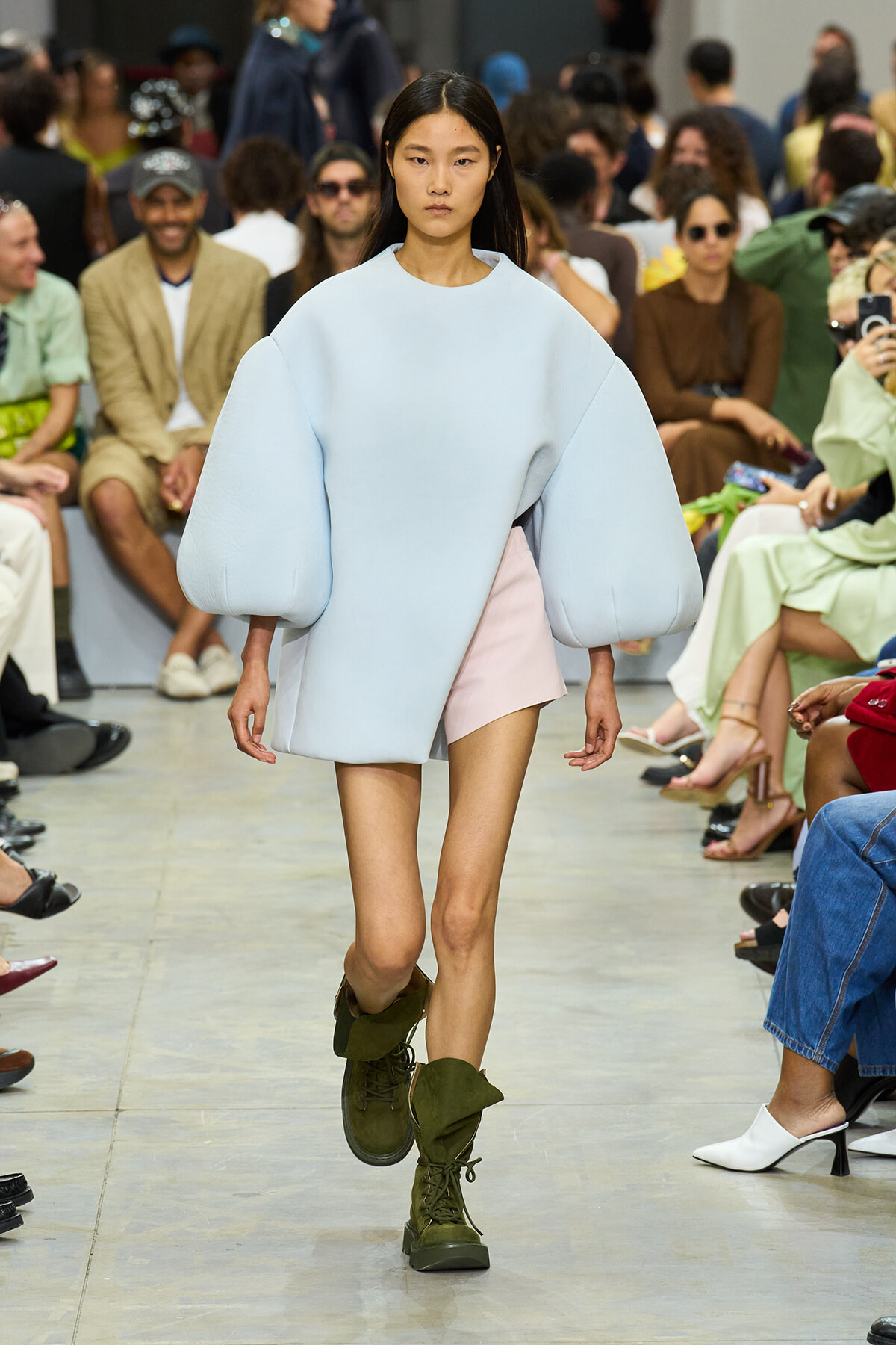 Model walks the runway in a pale blue oversized top with voluminous sleeves, paired with a pale pink mini skirt and olive green combat boots, audience in background.