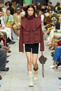 Model walks a runway in a burgundy oversized jacket with bold shoulder panels, black shorts, cream boots, and a brown handbag.