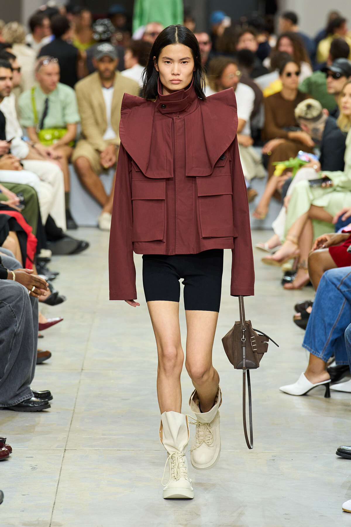 Model walks a runway in a burgundy oversized jacket with bold shoulder panels, black shorts, cream boots, and a brown handbag.