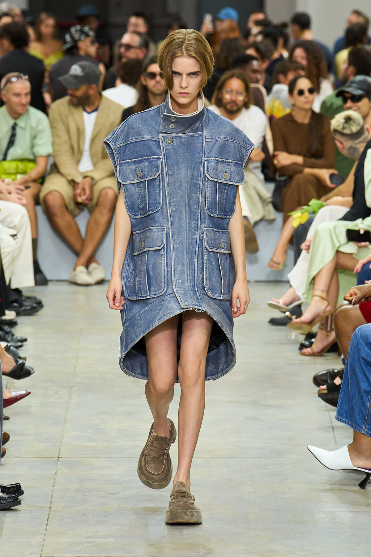 Model walks the runway in a sleeveless denim tunic with multiple front pockets and a high-low hem, as audience watches in the background.
